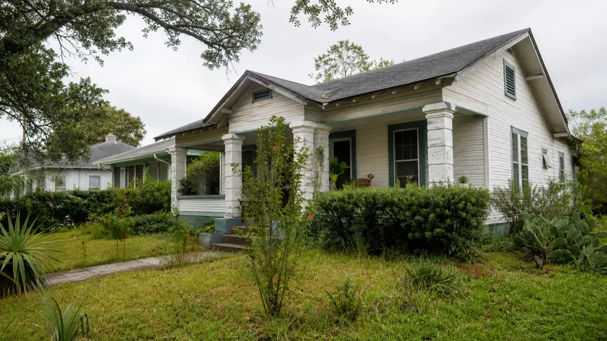 An older single-family home in South Texas with weathered paint and a slightly overgrown yard in soft daylight